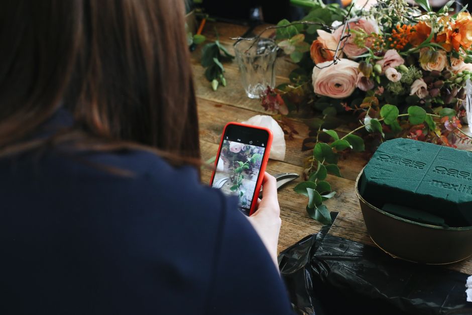 A person with brown hair wearing a dark blue top is taking a photo of a floral arrangement with a red smartphone. The arrangement consists of pastel pink and cream roses, orange blossoms, and green fo