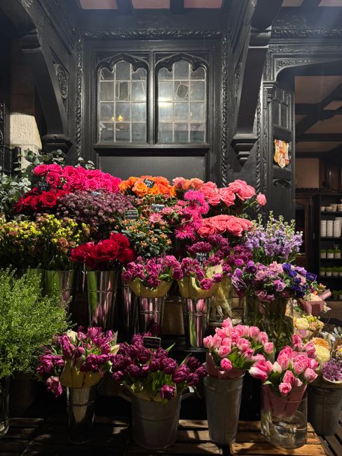 An indoor display of vibrant, fresh flowers arranged in clusters on a dark wooden shelf and in silver buckets, featuring pink, purple, red, and orange blooms including carnations and chrysanthemums. T