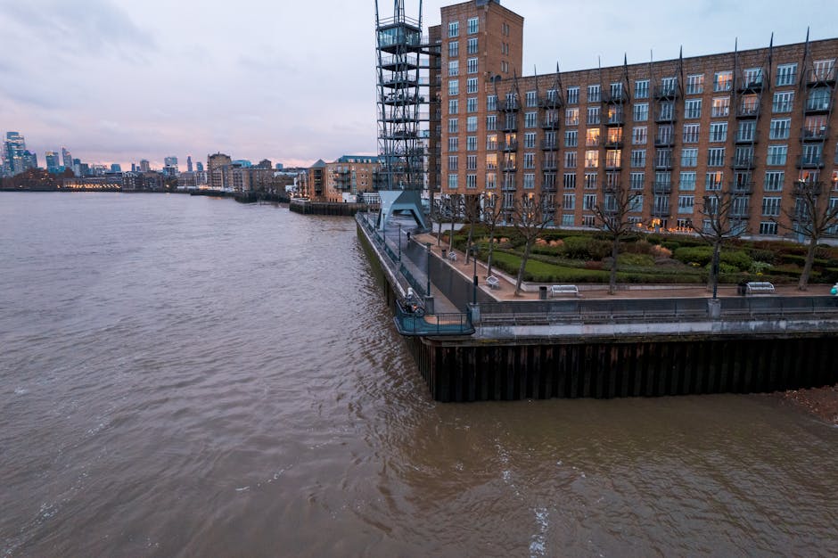 A scenic view of the Limehouse Basin featuring a modern brick apartment building with multiple windows and balconies, overlooking the river Thames. The foreground shows the water with gentle ripples, 