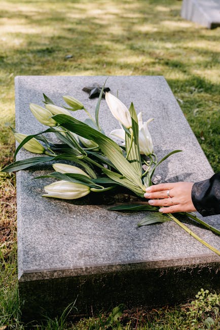 A person's hand, wearing a black leather jacket, gently placing a bouquet of white lilies and green foliage on a polished gray granite memorial plaque in an outdoor cemetery setting. The bouquet featu