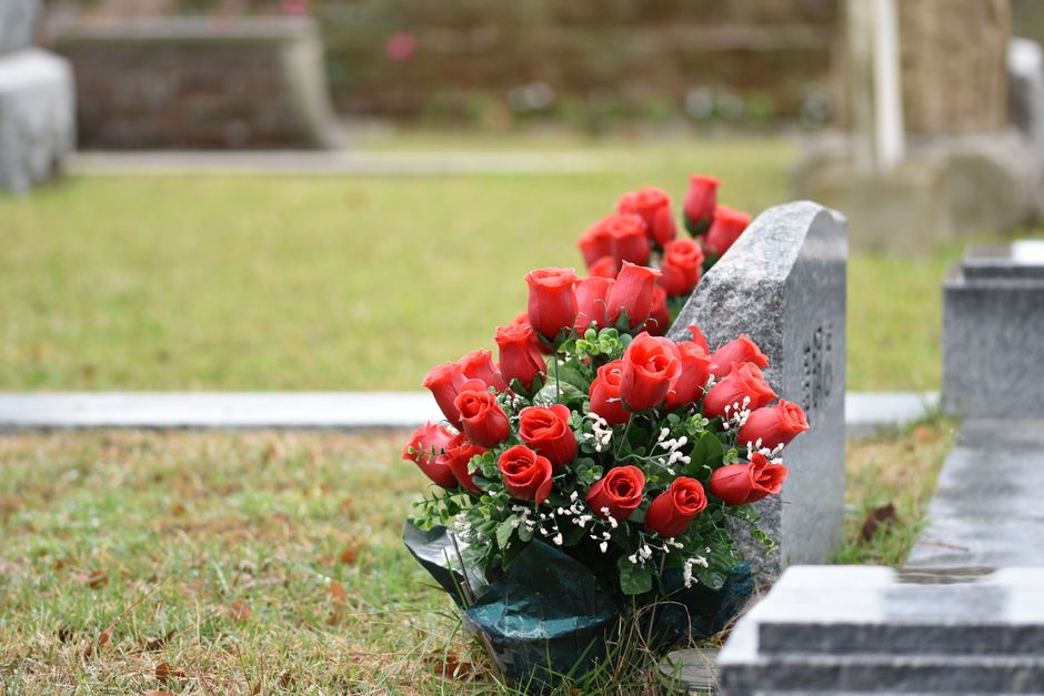 A bouquet of fresh red roses with tightly closed petals and green stems, arranged with small white filler flowers and dark green foliage, wrapped in black floral paper, resting against a gray stone gr