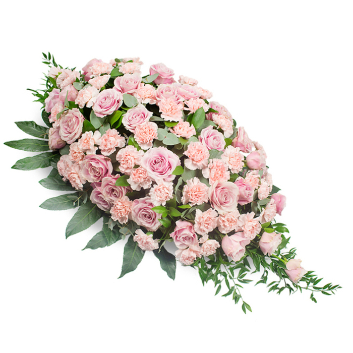 A long, tapering funeral spray of pink blooms lies against a clean white background, its shape forming a graceful diagonal teardrop that travels from a full, rounded upper section to a fine point at the lower right. The arrangement is richly layered with numerous pink roses, some fully opened to reveal complex whorls of petals, others still in bud or half-bloom, contributing a sense of depth and natural progression. Their colours range from very soft blush pink to a slightly deeper, dusty rose with hints of peach in the centres, creating a subtle play of tone that remains calm and soothing to the eye. Crowded between the roses, clusters of light pink carnations fill the design, their ruffled petals adding volume and a gentle, cushion-like effect. Together, the roses and carnations form a dense blanket of blooms that completely covers the underlying structure, suggesting meticulous craftsmanship and care. Around this core of flowers, dark green foliage shapes and supports the spray. Broad, oval leaves establish a firm outline, while finer, tapering sprigs of greenery extend discreetly beyond the flowers, particularly at the narrowing end, suggesting a soft, trailing motion. The lighting in the image is neutral and controlled, neither overly bright nor shadowed, allowing each detail to be clearly seen while maintaining a tranquil atmosphere. This arrangement feels like a thoughtful, respectful tribute, ideal as a casket spray or main sympathy piece for a funeral or memorial service in Limehouse, perhaps overlooking the water near Limehouse Basin or in one of the area's intimate chapels. It conveys quiet love, enduring memory, and a wish for peace, the kind of farewell local families might entrust to Flowers Limehouse when they want their sentiments expressed with elegance and restraint.