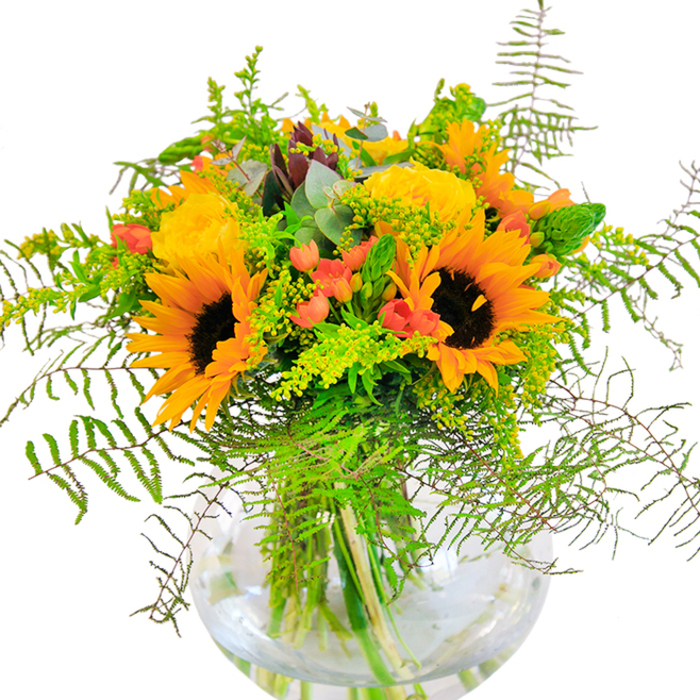 A first glance at this bouquet and the immediate impression is one of luminous celebration-a joyous composition echoing the riverside light of Limehouse Pier. Dominated by vibrant sunflowers with their signature yellow rays and deep, textured centers, the design is enriched further by tightly layered yellow garden roses, each bloom radiating gentle warmth. Interlaced with the centerpiece blooms, various shades of green-from elegant ferns to feathery solidago and slender, light-tipped spikes-spread outward, suggesting the resilient growth along the Thames Path. Accents of vivid red Hypericum berries dot the bouquet with their glossy vibrance, creating dramatic contrast against both blooms and greenery. Blue-green eucalyptus leaves add depth and rusticity, subtly softening the arrangement's contours. The entire ensemble is cradled in a clear, round glass vase, its transparency highlighting the clean lines and architecture of each submerged stem. View it closely, and you'll sense a harmonious balance between texture and colour, as well as a gentle blend of fragrances-floral, earthy, and fresh. Ideal as a gift for joyful occasions-a birthday or friendship celebration-or as a luminous highlight in any Limehouse kitchen or lounge, this expertly crafted bouquet brings a sense of local artistry and the natural brilliance of East London indoors.